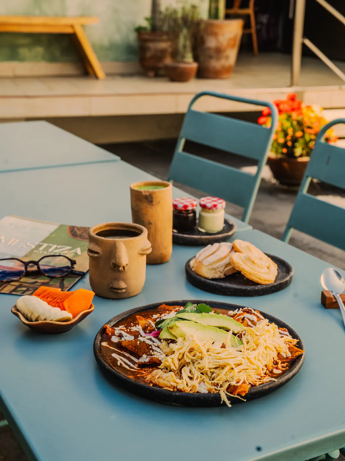 Mesa de desayuno en la terraza: platillo en barro negro, vasos de barro con rostros, conchas de pan y flores naranjas sobre una mesa turquesa