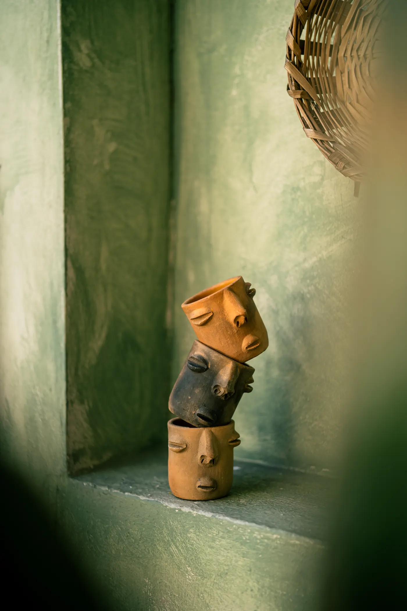 Vasos de barro con rostros prehispánicos apilados sobre un muro de estuco verde salvia
