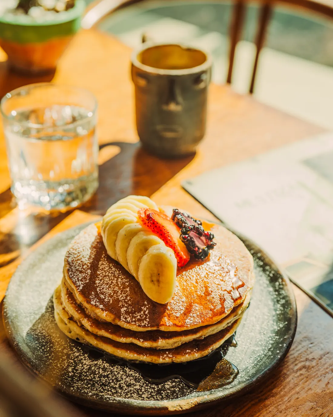 Hot cakes con rebanadas de plátano, fresa y azúcar glass, servidos en plato de cerámica sobre mesa de madera