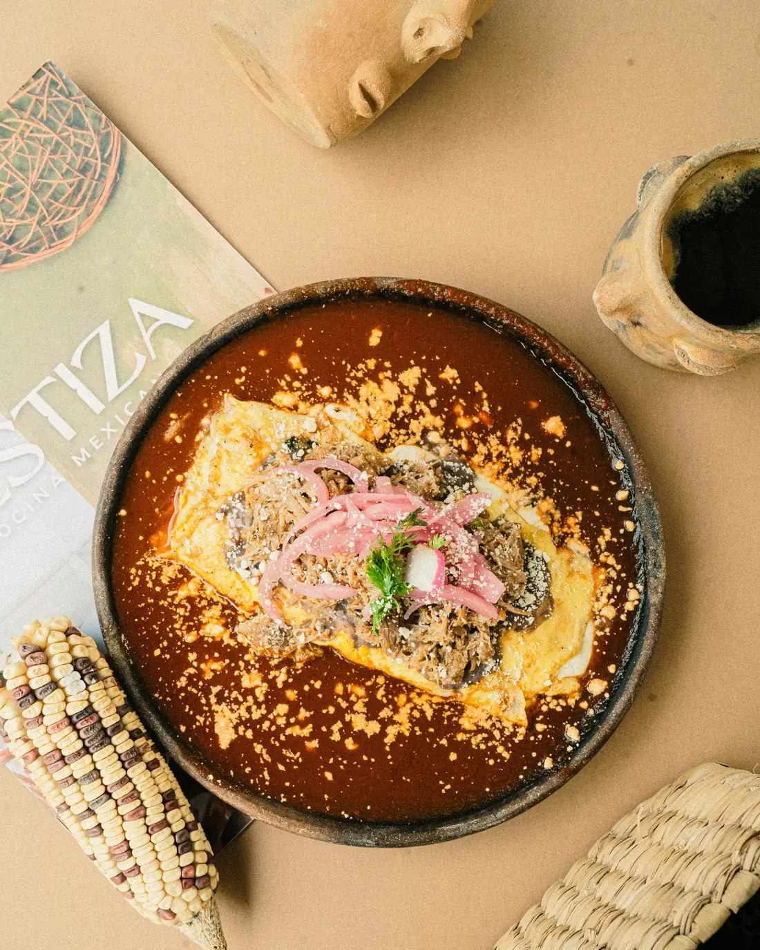 Omelette Mestizo relleno de cochinita pibil sobre salsa roja, con cebolla curtida y queso desmoronado, servido en plato de barro negro
