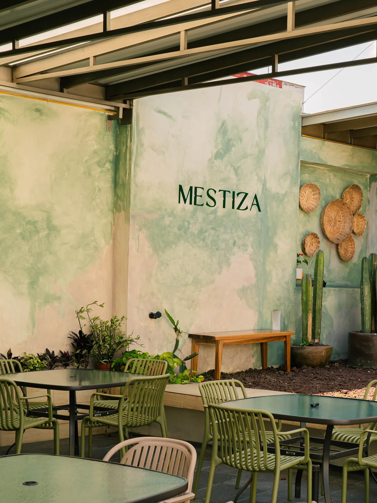 Patio de Mestiza: muro de estuco verde salvia con el nombre del restaurante pintado, tenates de palma colgados, cactus en macetas de barro y mesas del comedor al aire libre