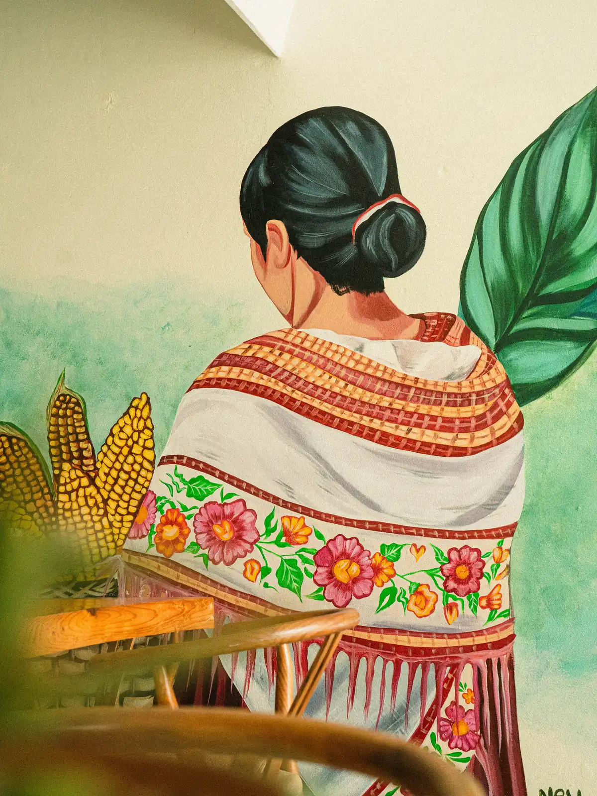 Mural de Mestiza: mujer de espaldas con huipil bordado de flores, entre mazorcas de maíz y hojas verdes